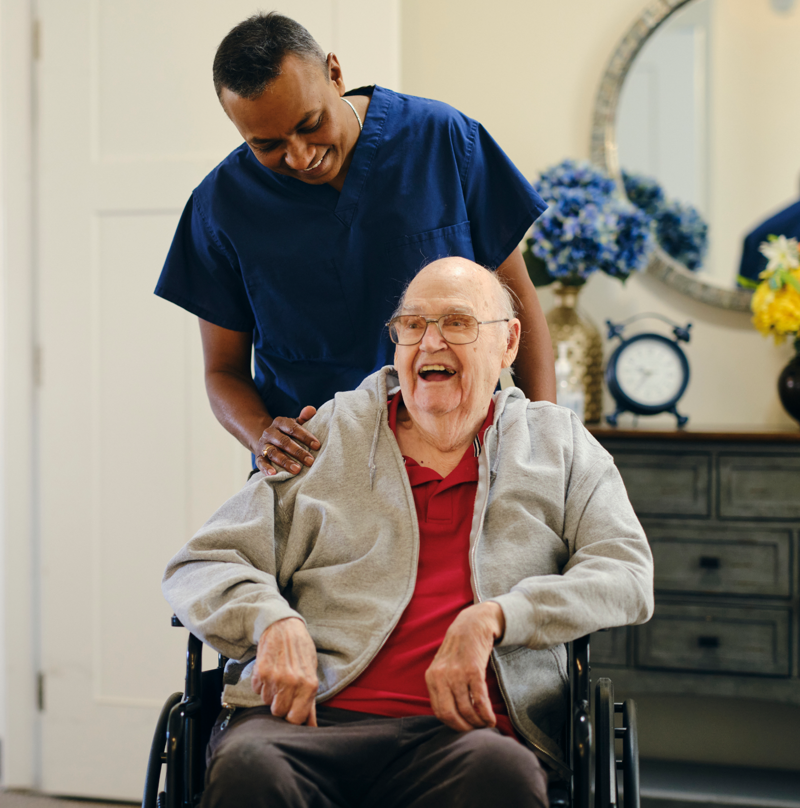 A heath care worker with his hand on the shoulder of a smiling elderly man in a wheelchair